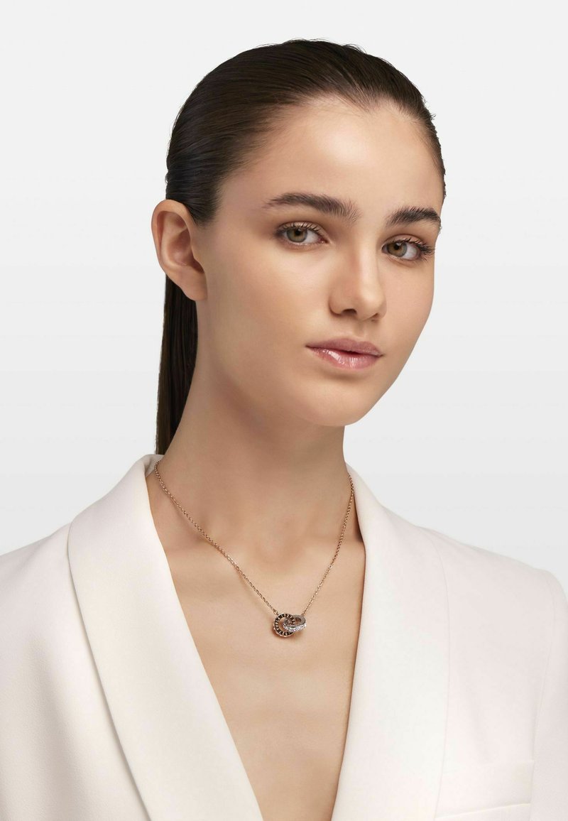 Rosen-guld halskæde med et sammenflettet cirkulært design med små klare ædelsten. Modellen bærer en hvid blazer, og håret er slicket tilbage.