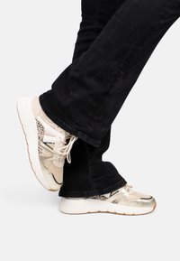 Baskets beiges et dorées avec des textures en maille et en cuir, arborant un accent en imprimé léopard et des lacets blancs, portées avec un pantalon noir.