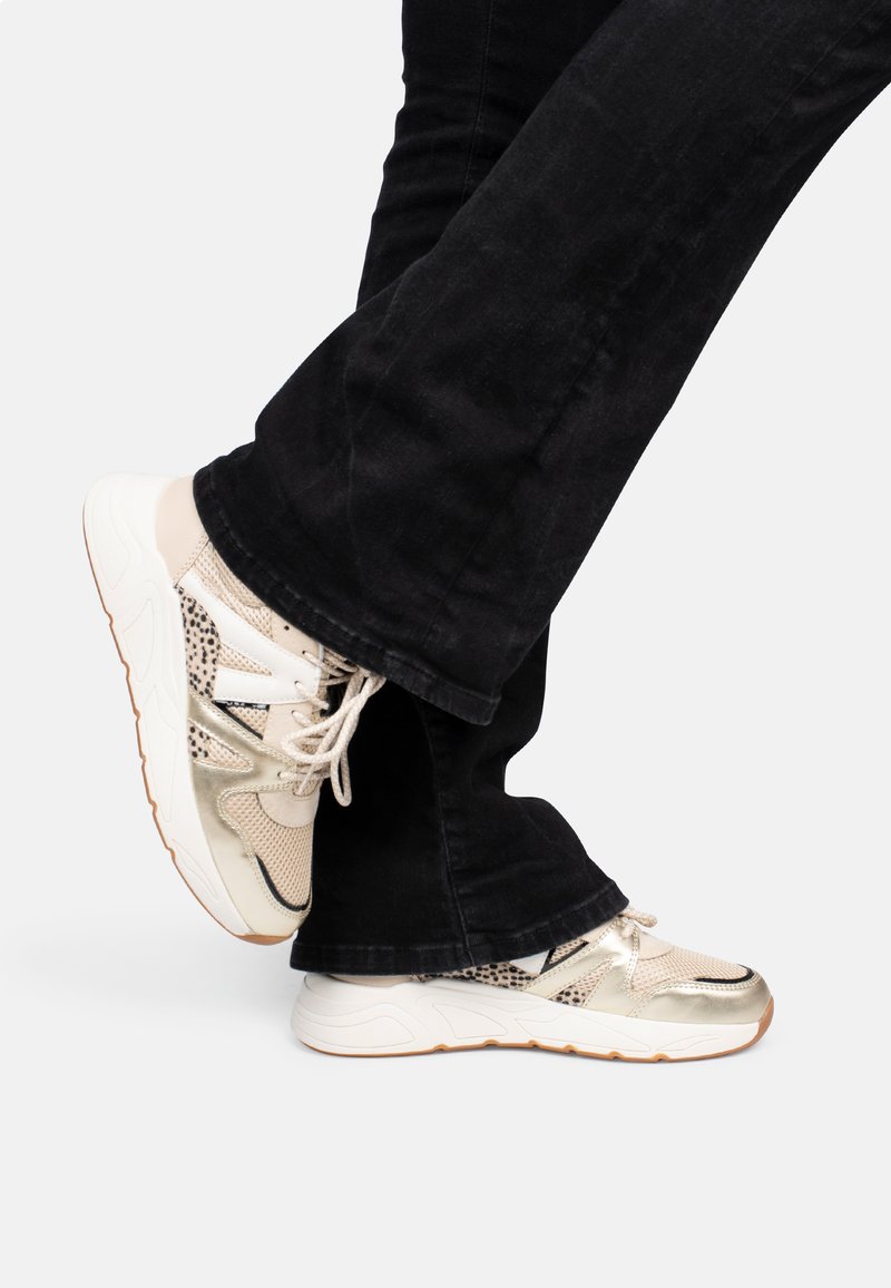 Baskets beiges et dorées avec des textures en maille et en cuir, arborant un accent en imprimé léopard et des lacets blancs, portées avec un pantalon noir.