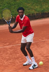 Polo shirt rossa con colletto bianco, pantaloni corti bianchi e scarpe Nike bianche con dettagli verdi. In mano tiene una racchetta da tennis su un campo in terra rossa.