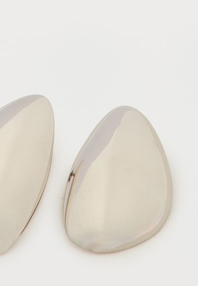 Két sima, polírozott fém cipőbetét lekerekített háromszög alakban, fehér háttér előtt.