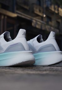 Ζεύγος λευκών παπουτσιών Adidas Ultraboost με ανοιχτό μπλε μεσαίες σόλες σε επιφάνεια από σκυρόδεμα, αστικό φόντο θολό.