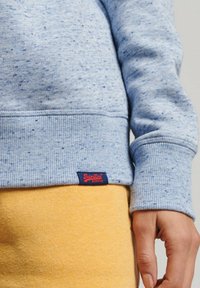Persona che indossa una felpa a pois blu chiaro e pantaloni gialli, con la mano destra rilassata lungo il fianco.