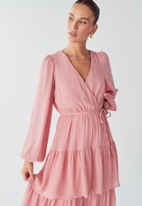 Růžové šaty s dlouhým rukávem, které mají hluboký výstřih do V, nařasený pas a vrstvené úpravy. Vyrobeno z lehké texturované látky.