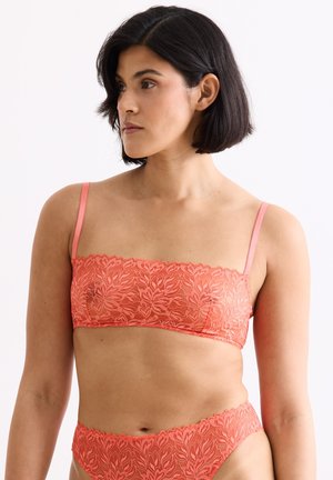 Frau mit kurzen dunklen Haaren, die ein korallenfarbenes Spitzen-Bandeau-BH und passende Slips trägt, schaut zur Seite vor einem einfarbigen Hintergrund.