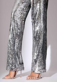 Srebrne spodnie z cekinami o błyszczącej, refleksyjnej powierzchni, z prostą nogawką i subtelnym rozszerzeniem przy kostce. Połączone ze srebrnymi sandałami na obcasie.