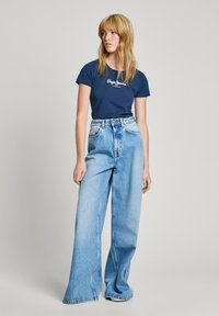 T-shirt marine avec logo blanc, associé à un jean en denim léger à jambes larges. Texture lisse, design taille haute et coupe longue.