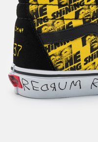 Sapatilha alta preta com tecido amarelo brilhante e gráficos de "The Shining". Acentuada com detalhes em vermelho e cinza, incluindo o texto "REDRUM".