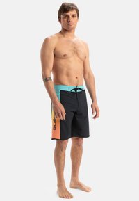 Bañadores de hombre en negro con una banda en turquoise. Cuenta con paneles laterales en naranja y amarillo con estampado de logo. Material ligero y de secado rápido.