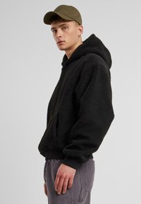 Sweat à capuche noir en polaire avec une poche sur le devant, capuche ajustable et poignets à côtes. Porté avec un pantalon en velours côtelé gris et une casquette verte.