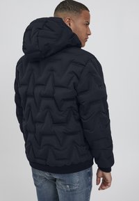 Svart pufferjakke med en teksturert sikksakk-mønster, hette og ribbede mansjetter. Båret over lyseblå slitte jeans, sett bakfra.
