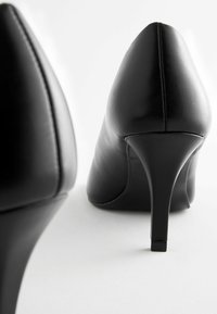 Black high-heeled shoes made of smooth leather with a pointed toe and slim stiletto heel, displayed from a rear angle against a light backdrop.