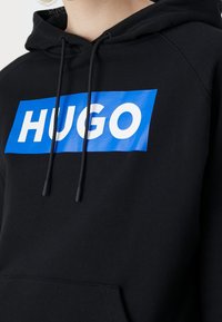 Sudadera negra con un destacado logo rectangular azul que presenta "HUGO" en grandes letras blancas. Tejido suave con cordones ajustables.
