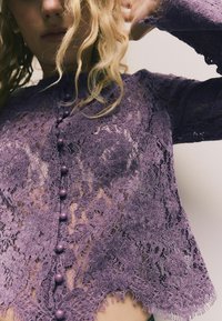 Lila Spitzenbluse mit floralen Mustern, einer gewellten Säume und runden Knöpfen an der Vorderseite. Lange Ärmel mit zarten Kanten.