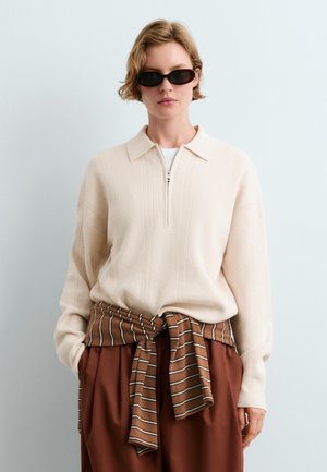 Donna con occhiali da sole neri, maglione di lana crema con cerniera frontale, pantaloni color ruggine e camicia marrone a righe legata in vita.