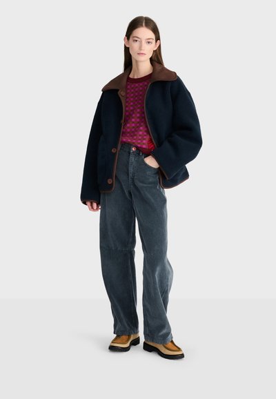Jeune femme debout portant une veste oversize bleu marine foncé, un pull violet à motifs, un pantalon large en velours côtelé gris et des bottes camel avec des semelles noires.
