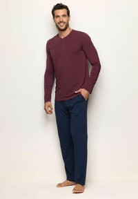 Man står barfota klädd i en vinröd långärmad henley och marinblå pyjamasbyxor mot en neutral bakgrund.