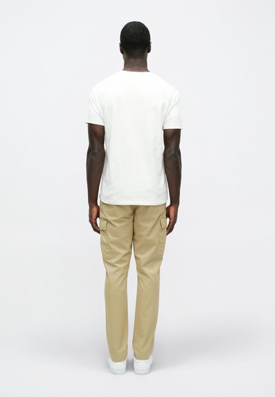 Λευκό βαμβακερό μπλουζάκι και μπεζ παντελόνι cargo, με πλαϊνές τσέπες, σε συνδυασμό με λευκά αθλητικά παπούτσια, εικονισμένα από πίσω σε απλό φόντο.