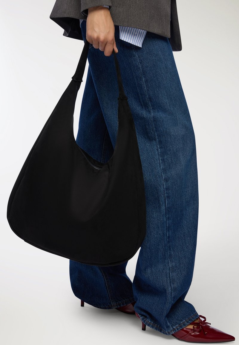 Borsa a spalla in tessuto nero con forma curvata e morbida al tatto, dotata di una sola tracolla, indossata insieme a jeans a gamba larga blu.