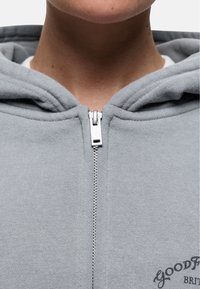 Felpa grigia con zip realizzata in tessuto morbido. Presenta una zip argentata, un cappuccio con cordino e testo stampato vicino all'orlo.