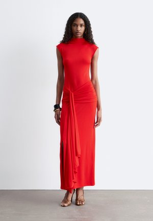 Kobieta z kręconymi włosami ubrana w czerwoną maxi sukienkę bez rękawów z bocznym rozcięciem i drapowanym detalem, stojąca na szarej podłodze na tle białej ściany.