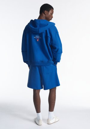 Homme portant un sweat à capuche bleu et un short assorti jusqu'aux genoux, avec des chaussettes blanches et des baskets, debout, les mains dans les poches, de profil.