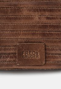 Morceau de cuir marron avec des lignes cousues, comportant une étiquette rectangulaire en relief avec "FREDs BRUDER". Surface texturée avec un aspect usé.