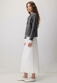 Ženska z dolgimi kodrastimi lasmi, oblečena v temno sivo oblikovano jopico, dolgo belo krilo in bele superge, stoji pred enobarvno ozadjem.