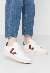 Zapatillas de piel blanca con logo rojo en forma de "V", detalles en el talón de color azul marino y cordones blancos. Suela texturizada con un patrón de dibujo visible. Combinadas con jeans azules.