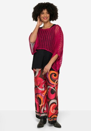 Rosa gehäkeltes Oberteil über einem schwarzen ärmellosen Shirt, kombiniert mit bunten, gemusterten weit geschnittenen Hose mit Wirbeln in Rot, Orange und Braun.
