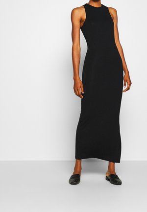 Mujer con vestido negro sin mangas hasta el tobillo, con cuello alto y zapatos negros sin cierre, de pie sobre un fondo liso.