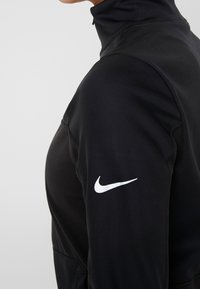 Sort Nike atletisk top med høj krave, tætsiddende design, lange ærmer og en hvid logo på højre skulder. Glat tekstur.