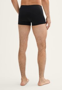 Zwarte katoenen boxershorts met een strakke pasvorm, voorzien van een gladde tailleband en minimale stiksels. Zichtbare gespierde benen en blote voeten.