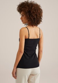 Frau mit lockigem Haar, die ein schwarzes, ärmelloses Oberteil und helle beigen Hosen trägt, von hinten vor einem schlichten Hintergrund gezeigt.