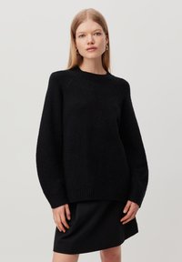 Black knit sweater with a round neckline and long sleeves. Ribbed hem and plush texture. Worn over a black skirt, model stands with hands relaxed.