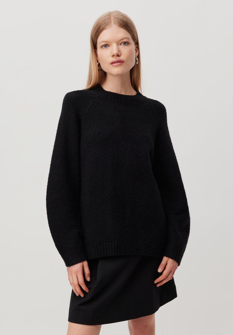 Black knit sweater with a round neckline and long sleeves. Ribbed hem and plush texture. Worn over a black skirt, model stands with hands relaxed.