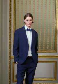 Σκούρο μπλε κοστούμι με ανάγλυφη πέτα, σε συνδυασμό με άσπρη πουκάμισο και μοτίβο μπλε χ bow tie. Απλός, κομψά ραμμένος σχεδιασμός. Χρυσά πλαίσια στο φόντο.