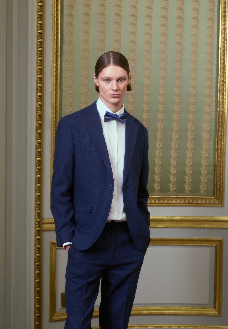 Σκούρο μπλε κοστούμι με ανάγλυφη πέτα, σε συνδυασμό με άσπρη πουκάμισο και μοτίβο μπλε χ bow tie. Απλός, κομψά ραμμένος σχεδιασμός. Χρυσά πλαίσια στο φόντο.