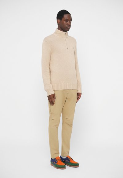 Beige kötött pulóver magas gallérral, világos bézs nadrággal párosítva, valamint színes, többféle textúrájú cipőkkel, amelyek zöld, narancs és kék részletekkel rendelkeznek.