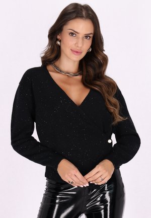 Schwarzer Wickelpullover mit tiefem V-Ausschnitt, verziert mit glitzernden Akzenten, langen Ärmeln und einem dekorativen Knopf. Kombiniert mit glänzenden schwarzen Hosen.