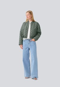 Padded zelena bomber jakna z okrogłym ovratnikom in rebrastimi manšetami, v kombinaciji s svetlobluznimi širokimi hlačami in odprto obuvalo. Gladka tekstura.
