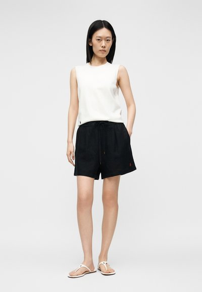 Γυναίκα στέκεται φορώντας λευκό αμάνικο τοπ, μαύρα σορτς με κορδόνι και λευκά σανδάλια σε απλό ανοιχτόφωτο φόντο.