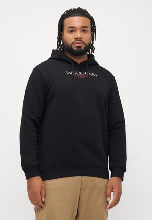 Muškarac s dreadlocksima nosi crnu Jack & Jones duksericu i bež hlače, stoji naslonjen na jednostavnu bijelu pozadinu.