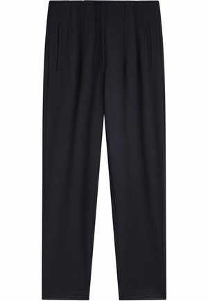 Personne portant un pantalon noir taille haute fuselé, un haut à manches longues gris foncé et des chaussures noires sans lacets, debout sur un sol lisse contre un mur blanc.