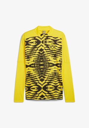 Gelbes langärmeliges Sportshirt mit einem halben Reißverschluss am Kragen, das ein auffälliges schwarzes Zickzackmuster und ein kreisförmiges Logo auf der Brust aufweist.