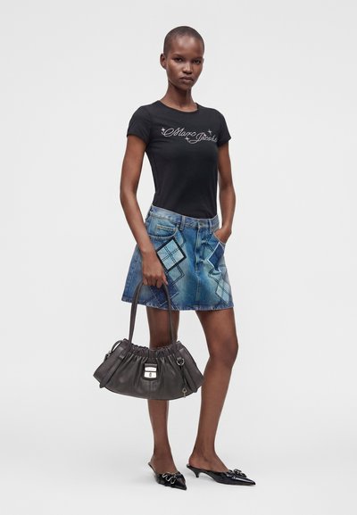 Γυναίκα με μαύρο μπλουζάκι Marc Jacobs, μπλε τζιν μίνι φούστα με λεπτομέρειες από patches, μαύρα μυτερά παπούτσια, κρατώντας μια μαύρη τσάντα.