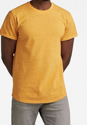 Gelbes Kurzarm-T-Shirt aus strukturiertem Stoff, mit rundem Halsausschnitt und lockerer Passform. Kombiniert mit grauen Jeans.