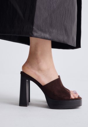 Pie que lleva un mule de ante marrón con puntera abierta y tacón alto, combinado con pantalones anchos oscuros y texturizados sobre un fondo liso y claro.