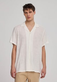 Camicia bianca di lino a maniche corte con colletto a punta, bottoni frontali e un motivo testurizzato. Indossata con pantaloni beige.