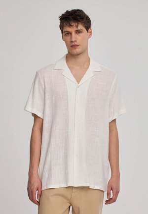 Camicia bianca di lino a maniche corte con colletto a punta, bottoni frontali e un motivo testurizzato. Indossata con pantaloni beige.